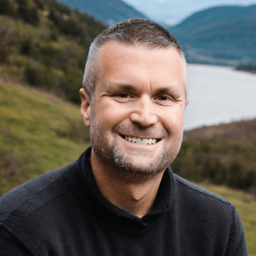 Beau wearing a black collared shirt sitting outside in a mountainous valley next to a lake.