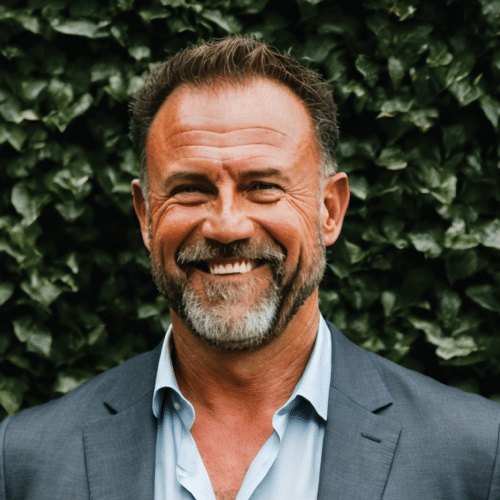 Greg smiling wearing a grey suit standing in front of a wall of some foliage.